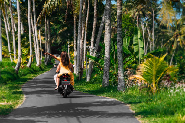 A happy young couple rides a scooter among the palm trees. A beautiful young couple is traveling on a scooter in Sri Lanka. Cheerful couple riding scooter at countryside road, vacation concept. stock photo