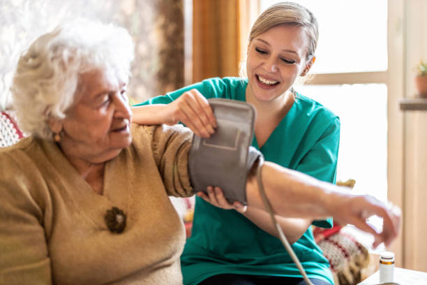 cuidadora femenina que mide la presión arterial de la mujer mayor en casa - asistente sanitario fotos fotografías e imágenes de stock