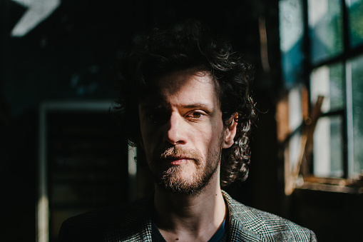 Portrait of a bearded man with curly hair standing in a dark room while direct sunlight exposes half of his face