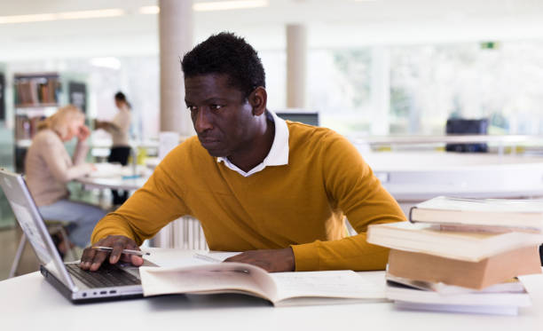 istock Efficient man preparing for exam in public library, reading book and using laptop for searching information 1307437993