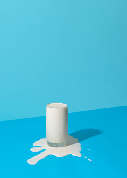 Overflowing glass of milk isolated on a blue background. Glass of milk overflowing on a blue background in bright light. Spilled milk puddle and glass of milk on a colored table. glass of spilled milk stock pictures, royalty-free photos & images