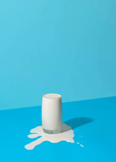 Glass of milk overflowing on a blue background in bright light. Spilled milk puddle and glass of milk on a colored table.