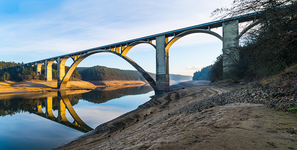 Beautiful sunny panorama with arched viaduct across riverbed, blue sky reflection in water surface and brown muddy bank. Podolsko, Czechia