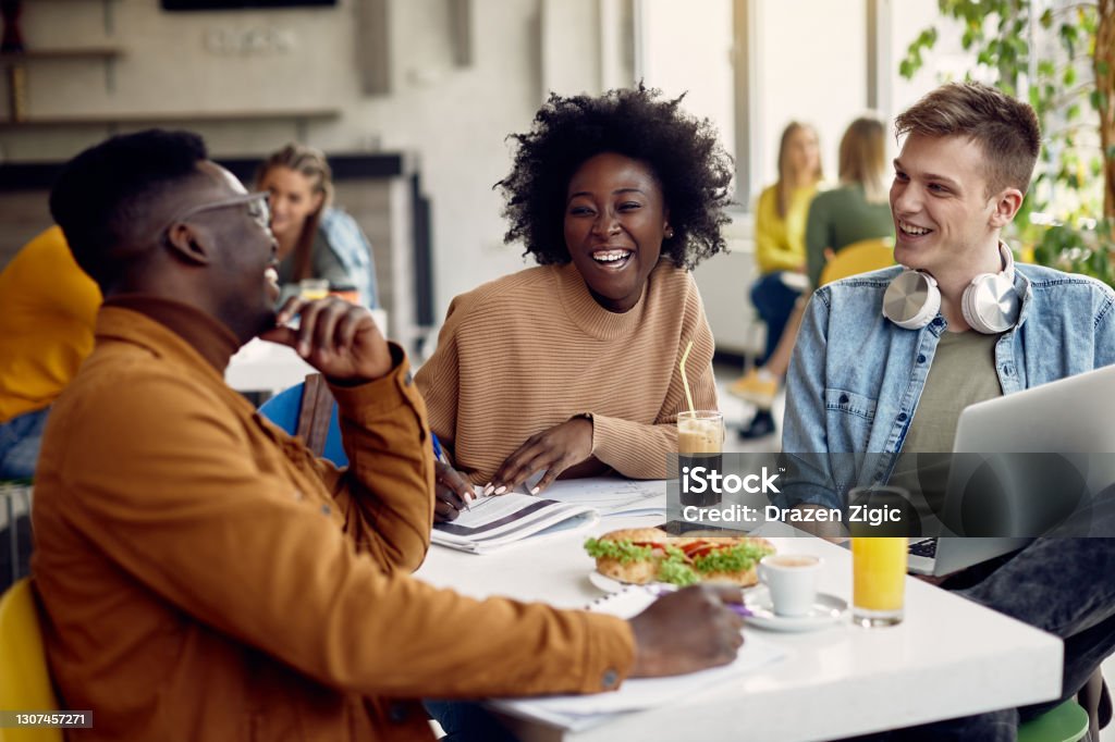 Cheerful group of college students having fun on lunch break in cafeteria. Group of happy students talking while studying at university cafeteria. Focus is on black female student. Cafeteria Stock Photo