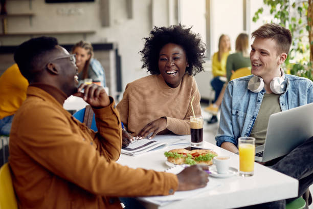 Cheerful group of college students having fun on lunch break in cafeteria. Group of happy students talking while studying at university cafeteria. Focus is on black female student. canteen stock pictures, royalty-free photos & images
