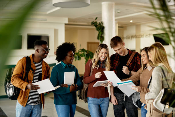 Group of happy students looking at exam results while standing at university hallway. Happy female student showing test results to her friends while standing in a lobby. public building stock pictures, royalty-free photos & images