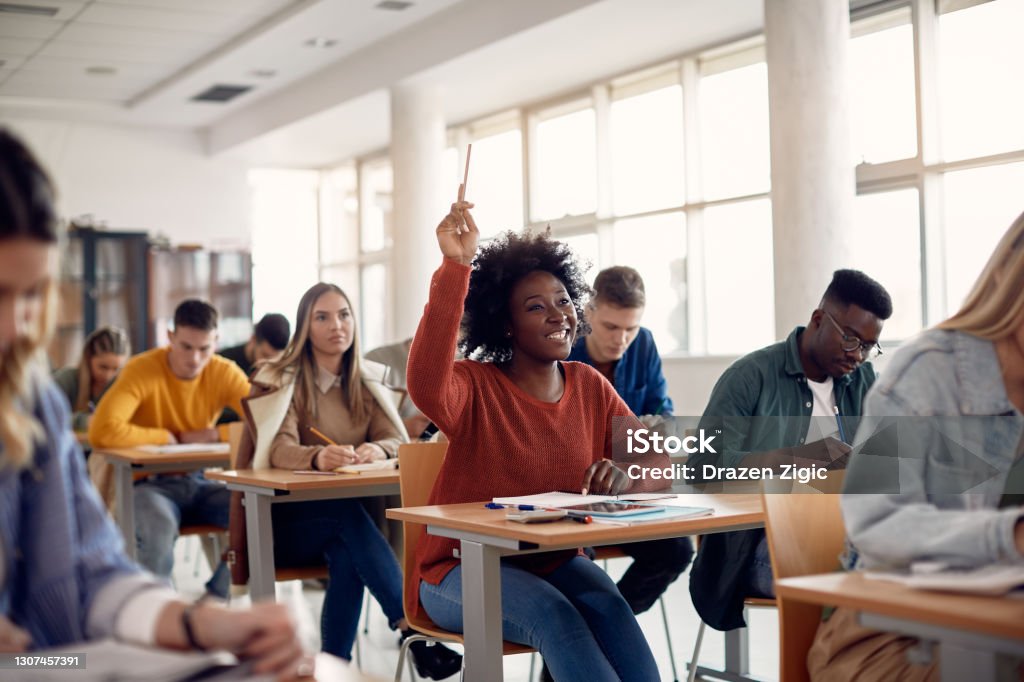 Happy black student raising arm to answer question while attending class with her university colleagues. Happy African American student raising her hand to ask a question during lecture in the classroom. Classroom Stock Photo