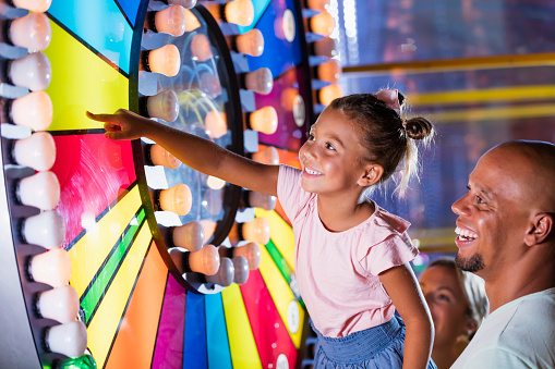 A 5 year old mixed race African-American and Caucasian girl having fun with her father at an amusement arcade. She is reaching up and touching a colorful, illuminated spinning wheel. Dad is a mid adult African-American man in his 30s. Mom is out of focus in the background.