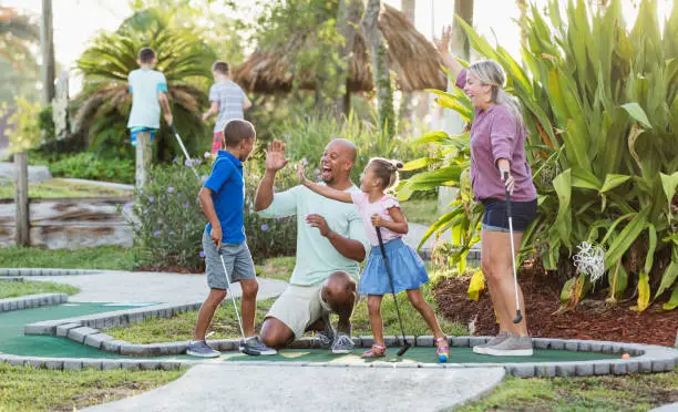 An interracial family having fun together playing miniature golf. The African-American father and Caucasian mother are in their 30s. The children are 9 and 5 years old. Dad and sister are giving the boy high fives for his winning putt.
