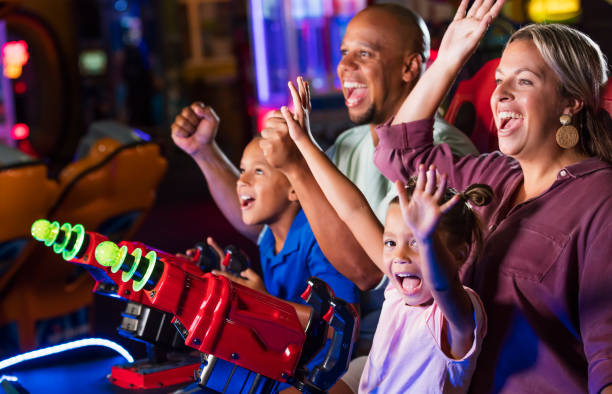 Mixed race family playing game at video arcade An interracial family having fun together playing at an amusement arcade. The African-American father and Caucasian mother are in their 30s. The 9 year old boy and 5 year old girl are sitting in their parents' laps playing a video arcade game. The main focus is on mother and daughter. Everyone is cheering after winning the game and the little girl is looking toward the camera. arcade stock pictures, royalty-free photos & images