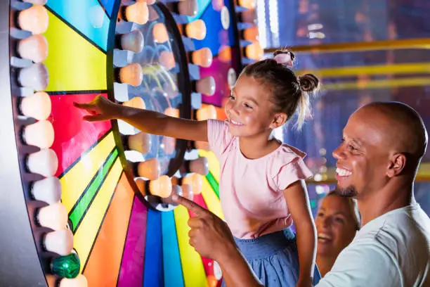 A 5 year old mixed race African-American and Caucasian girl having fun with her father at an amusement arcade. She is reaching up and touching a colorful, illuminated spinning wheel. Dad is a mid adult African-American man in his 30s. Mom is out of focus in the background.