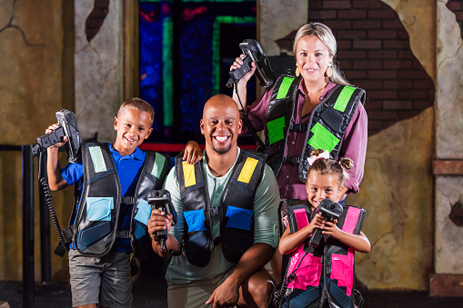 An interracial family having fun together at an amusement arcade playing laser tag. The African-American father and Caucasian mother are in their 30s. The two mixed race children are 9 and 5 years old. They are smiling and the little girl is aiming her laser toward the camera.