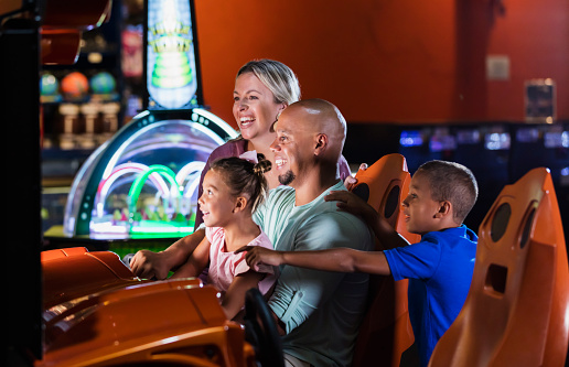 An interracial family having fun together playing at an amusement arcade. The African-American father and Caucasian mother are in their 30s. The children, and boy and girl, are 9 and 5 years old. They are smiling and watching as the daughter, sitting in dad's lap, is in the driver's seat, playing an arcade game.