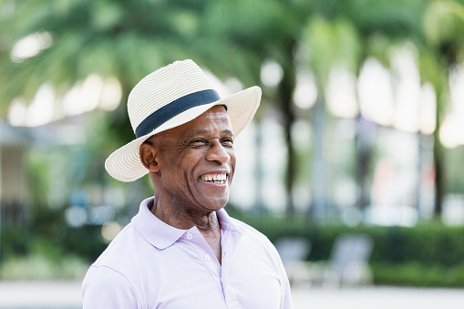 A senior African-American man in his 70s wearing a short sleeve collared shirt and wide brimmed hat, standing on the deck of a swimming pool, smiling confidently.