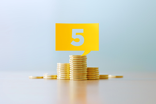 Number 5 written yellow speech bubble sitting over coins stacks before defocused background. Horizontal composition with copy space. Great use for budget concepts.