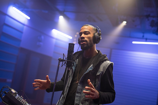 Young male singer with eyes closed gesturing while singing and recording songs in recording studio.