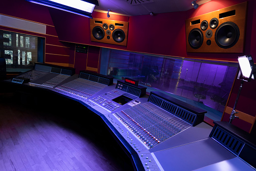 A serious black male audio engineer thinking while looking at a computer screen in a music studio. He has long curly hair and wears cool clothes. He is sitting inf front of an audio mixer with orange lights. There are several loudspeakers surrounding the sitting male. The lights in the studio are violet.