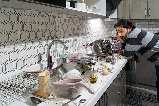 Erkekler evde yalnız kaldıklarında mutfak dağınık hale gelir. Mutfak tezgahında bekleyen kirli bulaşıklar, çöpe atılmamış atıklar. Bu dağınıklığa yorgun ve endişeli gözlerle bakan adam. full frame makine ile çekilmiştir