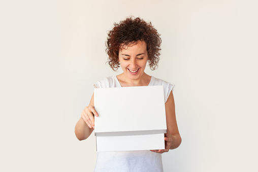 A curly-haired young woman opens a white shoe box that she ordered from an online store. The company delivers packages quickly to your home. Anniversary surprise. White background.