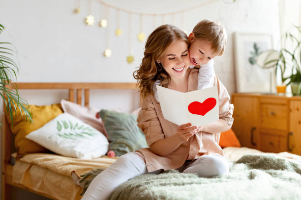 Mother and son reading greeting card Cheerful mother hugging son and reading handmade greeting card with heart while resting on bed during holiday celebration mothers day  at home mothers day stock pictures, royalty-free photos & images