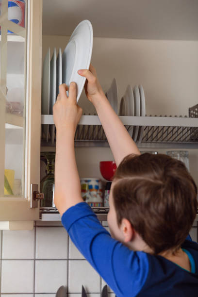 istock Kid boy sitting on the kitchen worktop and putting dishes in cupboard 1307469695