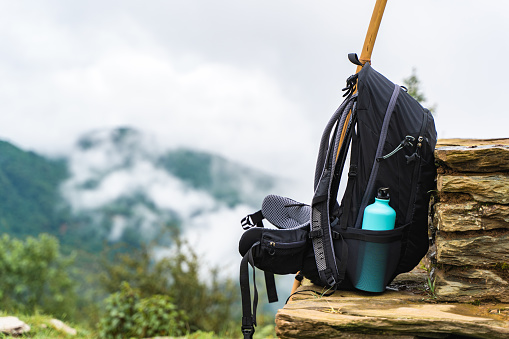 Backpack with water bottle and bamboo stick on stone fence near chasm. Mountains are hidden by clouds. Hiking and trekking concept.