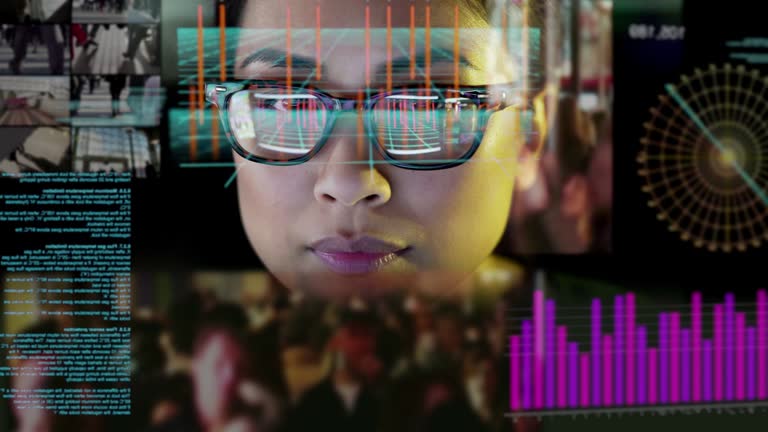 4K stock video clip of a young Chinese Asian woman sitting in a dark office. She is intently reading data from a virtual see through screen displaying moving text, graphs & diagrams which are reflected in her glasses.