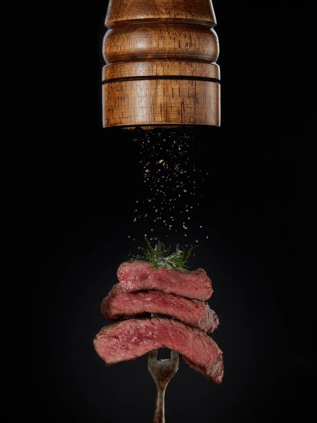 istock Steak menu. Milled spices falling from pepper mill on grilled pieces of beef steak medium rare on fork on black background. 1307483545