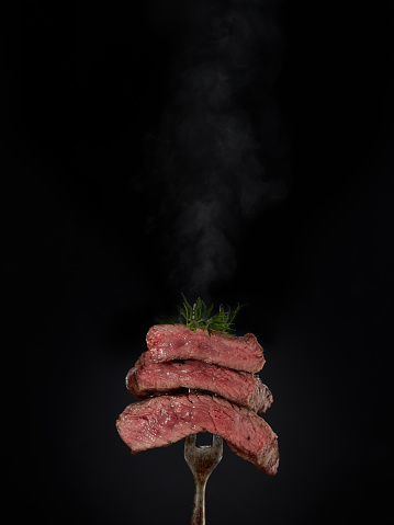 Roasted meat. Grilled hot pieces of beef steak medium rare with smoke on fork on black background.. Steak menu. Grilled menu.