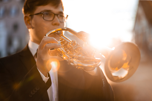 Trumpet Player in the City. Well-dressed young man in black suit with bow tie playing a trumpet in the city during sunset. Backlit from the sunset light. Small DOF, Selective Focus on Trumpet. Trumpet Player Millennial Generation Portrait.