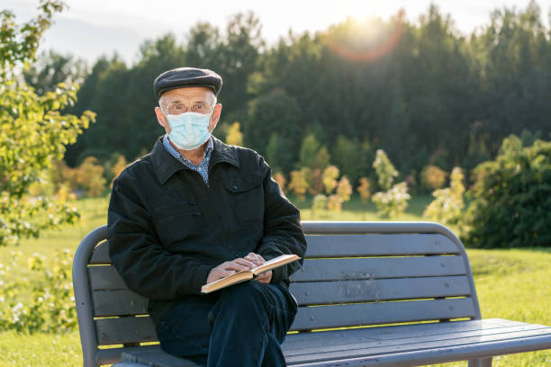 istock Senior man in cap and facemask sitting on bench and holding book 1307498342
