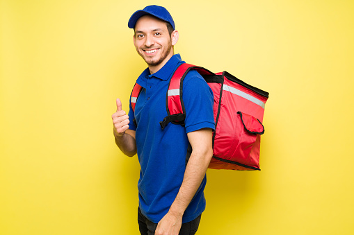 Attractive man standing against a bright yellow background while wearing a red delivery backpack. Male courier delivering takeout food for a delivery app