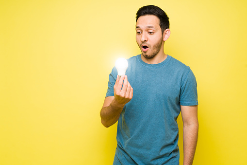 Handsome man holding a lighted light bulb and looking surprised. Shocked latin man in his 30s having a new creative and bright idea