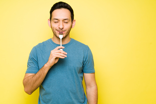 Hungry latin man with his eyes closed enjoying a spoonful of food. Handsome hispanic man eating delicious food and holding a fork in his mouth