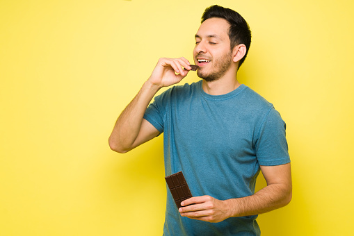 Attractive latin man eating a big chocolate candy bar. Handsome man in his 30s with sugar cravings enjoying a chocolate