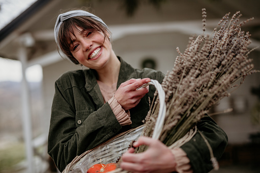 Portrait of a beautiful young adult woman with bangs, carrying a white wooden basket filled with Lavender flowers, loving the smell, looking at the camera with a big smile on her face, while enjoying the fresh air and her white wooden cabin house