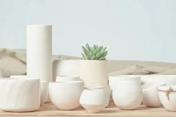 All kinds of handmade white clayware, randomly combined and placed on the unpainted wooden table. The light background environment is very bright, fresh, elegant and energetic. There is also a small green succulent plant for decoration.