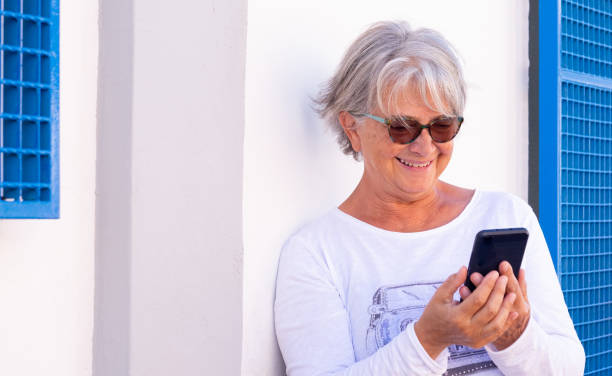 istock Senior woman smiles looking her smartphone. Standing against a white wall using wireless technology. Joyful elderly lifestyle 1307533218