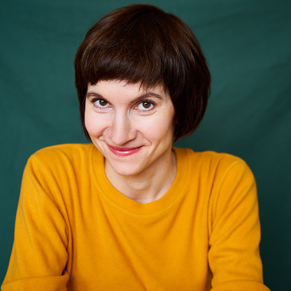 Smiling mature brunette woman looking playful and defiant, close-up face portrait of middle-aged lady in bright yellow sweater on bright colorful dark green textile background. Generation X, 40s