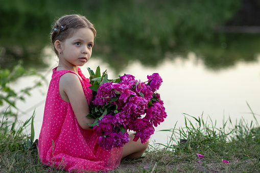 Little girl sitting with a bouquet of flowers on the shore of the pond