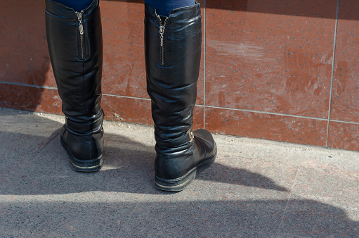 Women's legs in high leather boots. Black boots with metal zippers against the background of a red wall. Outside the premises. Daytime. Back view.