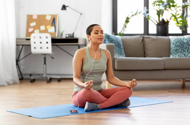 woman listening to music and meditating at tome mindfulness, spirituality and healthy lifestyle concept - african american woman in earphones listening to music on smartphone and meditating in lotus pose at yoga studio sukhasana stock pictures, royalty-free photos & images