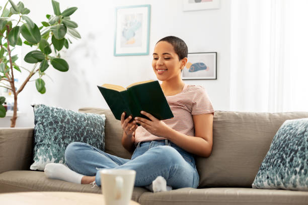 happy african american woman reading book at home people and leisure concept - happy smiling african american woman sitting on sofa and reading book at home reading stock pictures, royalty-free photos & images