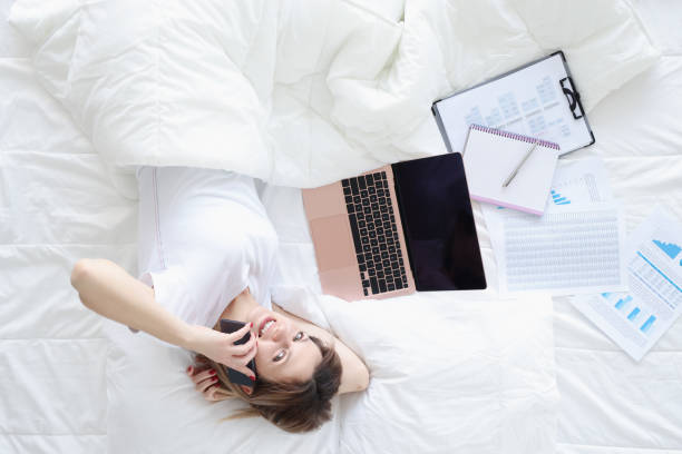 istock Smiling woman talking on phone and working on laptop while lying on white bed 1307543509