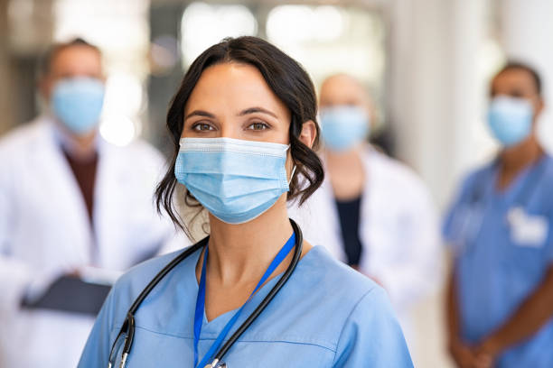 Happy nurse with face mask smiling at hospital Close up face of confident beautiful female nurse in front of his medical staff looking at camera while wearing protective face mask due to covid-19 virus. Smiling young surgeon standing in front of his healthcare colleagues wearing surgical mask. Portrait of satisfied and successful doctor with team in background in private clinic. nurse photos stock pictures, royalty-free photos & images