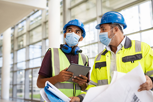 Mixed race architect and technician at construction site reviewing blueprints while wearing surgical face mask and hardhat. Team of specialists construction worker with face mask using digital tablet with plans. Young indian architect with mature engineer wearing hard helmet and protective mask for safety against coronavirus while working.