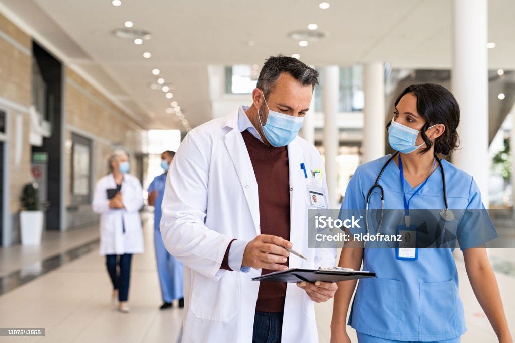 Mature doctor and nurse discussing patient case General practitioner and young nurse wearing surgical face mask against covid-19 while having a discussion in hospital hallway. Doctor with face mask discussing patient case status with his medical staff while walking on corridor. Worried busy doctor showing medical report to nurse and wearing protective face mask with copy space. Doctor Stock Photo