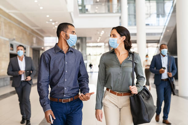 Businesspeople talking while walking with face mask Multiethnic business man and businesswoman walking with face mask looking at each other and talking at modern workspace. Two colleagues wearing protective mask due to covid-19 while going to work, new normal concept. Indian young man and latin woman discussing project while walking and keeping social distance. covid secure photos stock pictures, royalty-free photos & images