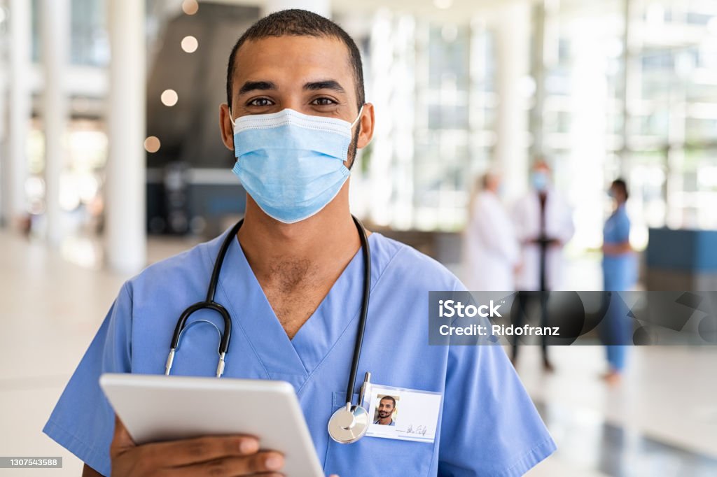 Middle eastern nurse wearing face mask at hospital Portrait of young multiethnic doctor using digital tablet wearing safety surgical face mask at private clinic during covid pandemic. Confident indian healthcare worker working on laptop and looking at camera. Successful smiling nurse wearing mask for safety against covid-19 with copy space. Nurse Stock Photo