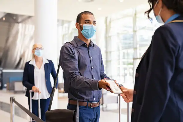 Indian passenger wearing surgical mask showing e-ticket to flight attendant at boarding gate. Young mixed race businessman showing boarding pass on mobile phone to air hostess while wearing protective face mask during covid pandemic. Multiethnic business man in a row with flight reservation hand the phoone to stewardess at airport keeping social distance.
